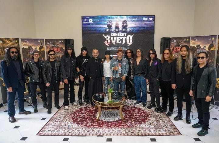 Group of musicians posing for a photo in a room with posters and a large screen showing a concert banner behind them.