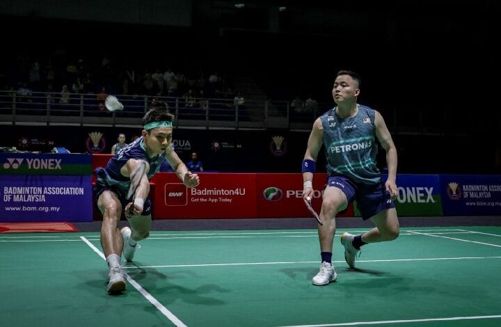 Two badminton players sprint toward the shuttlecock on a green court during a match, one in teal and blue reaching low for the shot while the other stands ready with a racket.