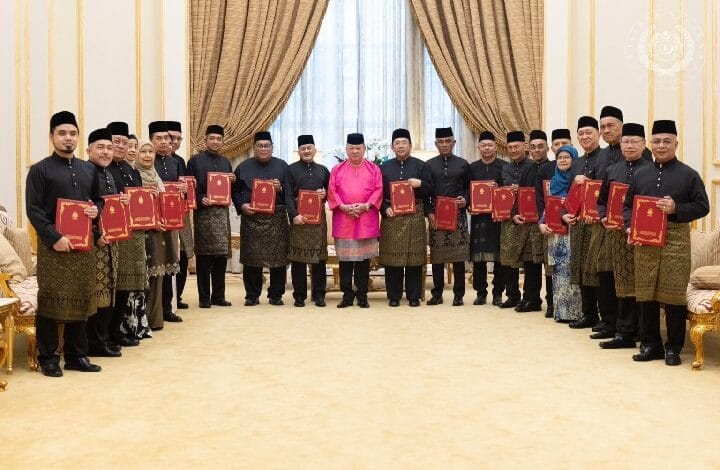 Group of people in traditional Malay attire pose with red certificates in a formal ceremony in a grand room with gold curtains.