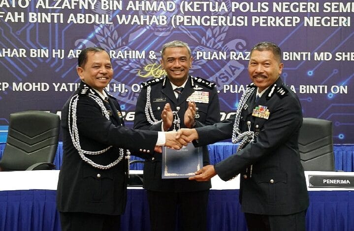 Three uniformed police officers on a stage shake hands while presenting a framed award flagship ceremony backdrop behind them