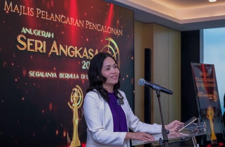 Woman in a white blazer and purple top speaks at a podium with a microphone during an awards ceremony, a large screen behind shows Ser i Angkasa branding.