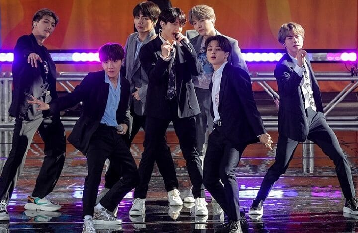 A seven-member boy band performing a dynamic dance on stage with neon purple lighting in the background, wearing suits and casual outfits.