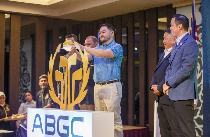 Man in a blue shirt lifts a gold circular trophy toward the audience on a stage with an ABGC sign; several people watch nearby in a formal setting.