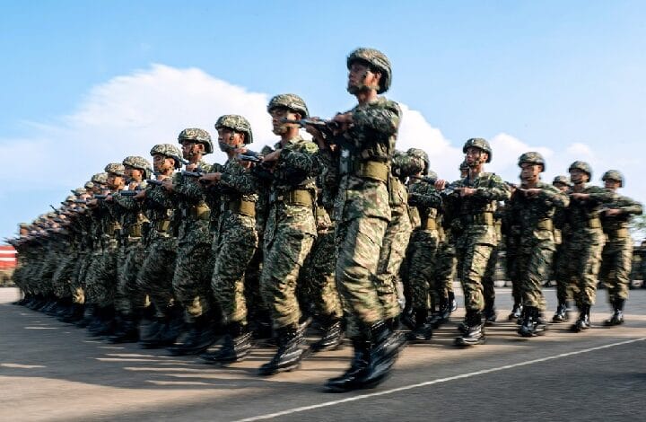 Row of soldiers in camouflage uniforms marching in formation, rifles raised, on a sunny parade ground