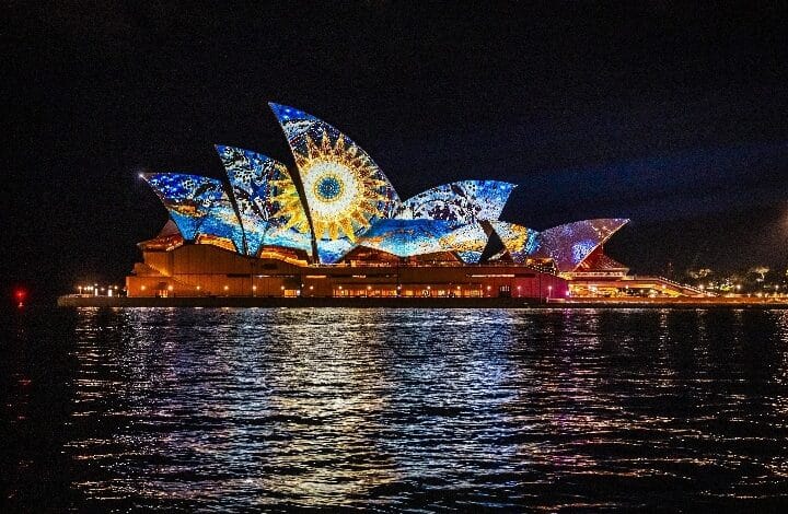 Sydney Opera House at night illuminated with a colorful projected light display reflecting on the harbor on calm water.