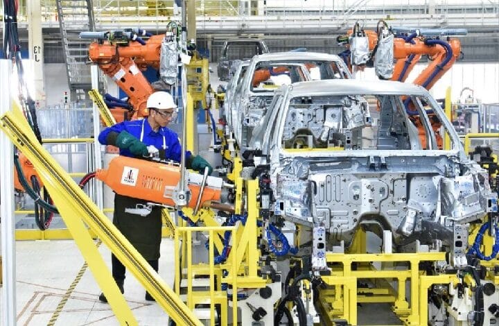 Factory assembly line: worker in a hard hat uses an orange robot arm beside a partially assembled silver car body on a yellow frame.