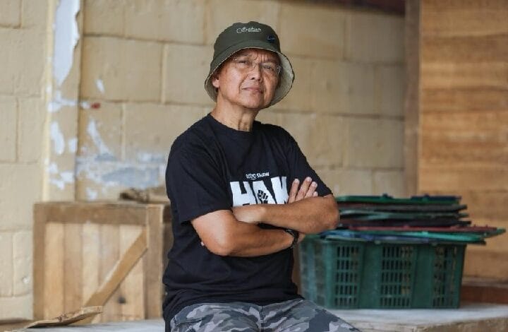 Older man wearing a bucket hat and black t-shirt with a white logo, standing with arms crossed in a workshop-like space with crates in the background.