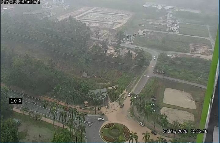 Aerial view of a roundabout with palm trees and roads; construction plots in the hazy background. Timestamp 23 May 2026 15:12:46.