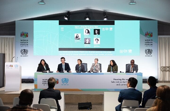 Panel of six speakers seated at a long table on a stage, with a large projection screen and World Urban Forum banners behind them.