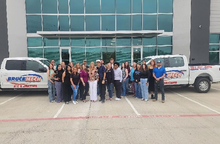 Group of about 25 people posing outside a modern glass-front office, flanked by two white service trucks with blue and red logos.
