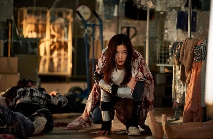 Young woman crawls through a debris-filled industrial space, bloodied clothing, looking at the camera.