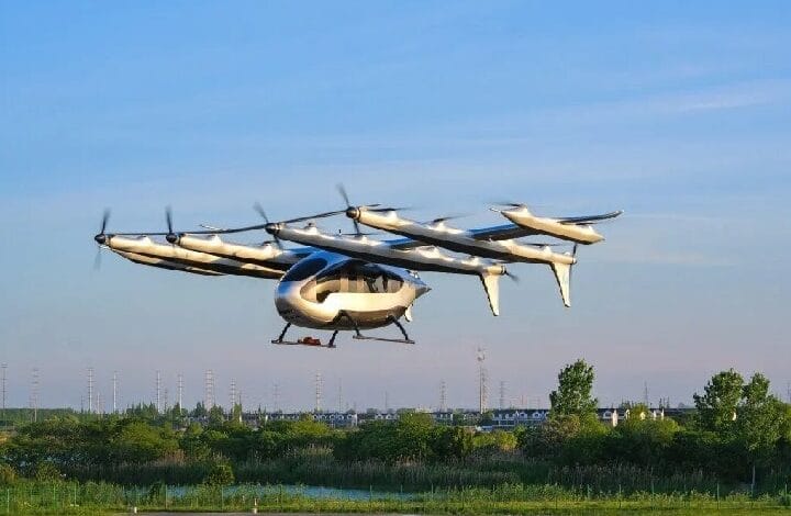 Six-rotor VTOL aircraft hovering above a grassy field with trees and power lines in the distance.