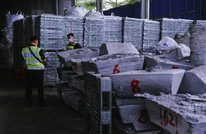 Two workers in reflective safety vests and masks stand amid stacks of large metal ingots in a warehouse.