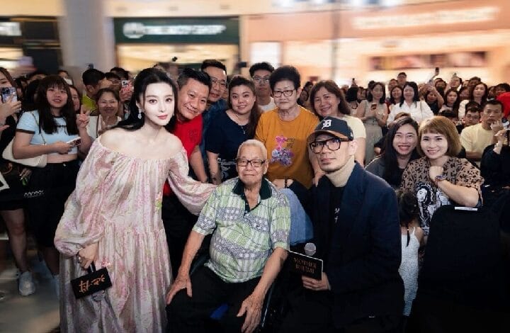 Diverse group of fans posing for a crowd photo at a public event; a young woman in a pink off‑the‑shoulder dress stands at the left, an elderly man in a wheelchair sits center, flanked by smiling fans in a busy venue.