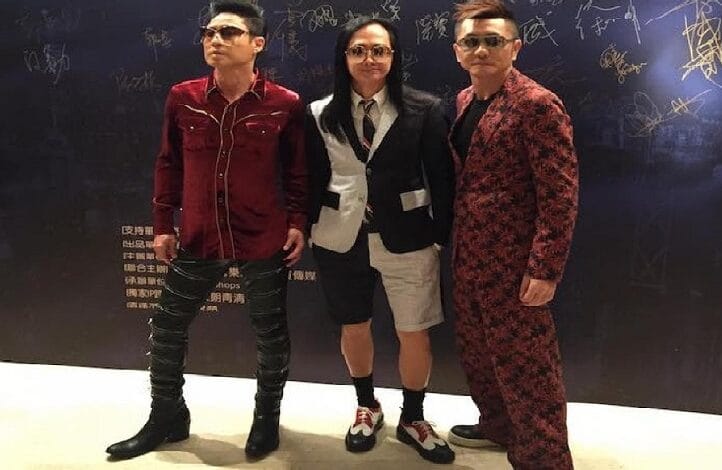 Three people wearing sunglasses pose indoors against a dark wall; left person in a red shirt with black pants, center person in a black-and-white blazer with tie and shorts, right person in a red patterned suit.