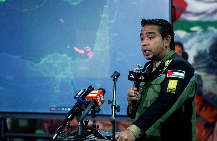 Man in a green camouflage-style jacket speaks at a press conference with multiple microphones, a map projected on a screen behind him.