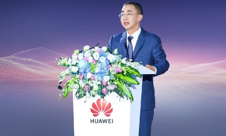 Man in a blue suit giving a speech at a podium with a Huawei logo and floral arrangement nearby.