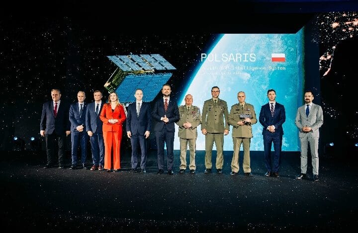 Group of ten people in business suits and military uniforms stand in a line on a stage, posing for a formal photo against a space-themed backdrop that displays the word POLSARIS and a small flag.