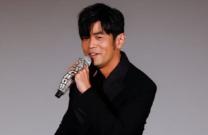 Male singer in a black suit holding a jeweled microphone during a performance on a plain gray background: midshot focus on the mic and face.