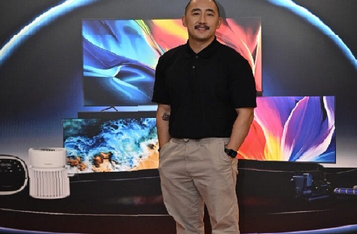 Man in a black polo and khaki pants stands with hands in pockets in front of a multi-panel, colorful TV display backdrop in a showroom.