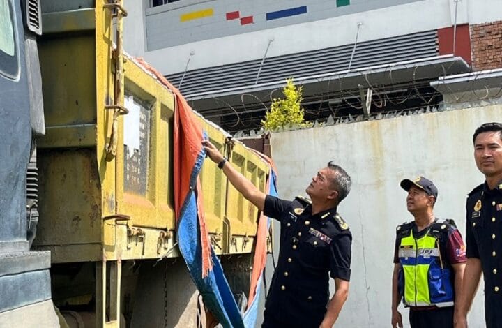 Three uniformed men inspect a yellow dump truck trailer; one points to a blue and orange tarp while two watch nearby in an outdoor area.