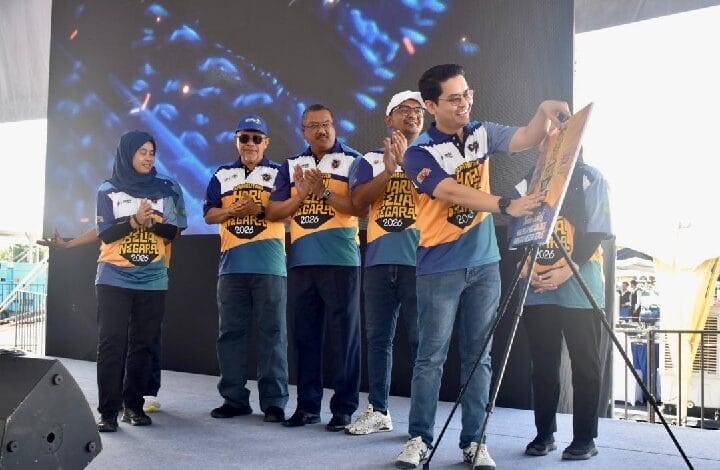 Six people on a stage in matching blue and yellow shirts; one man holds a poster while others clap underneath a large screen backdrop.