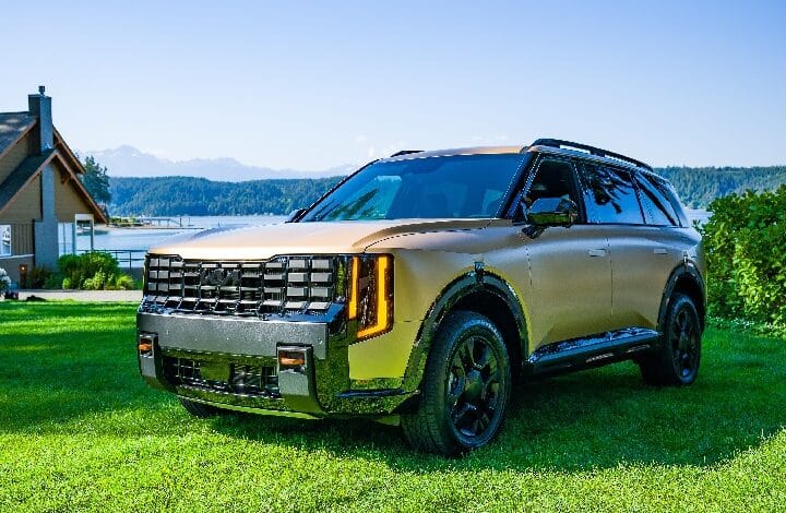 Gold-colored SUV parked on a grassy lawn by a lakeside property with a house and distant mountains in view, on a sunny day.