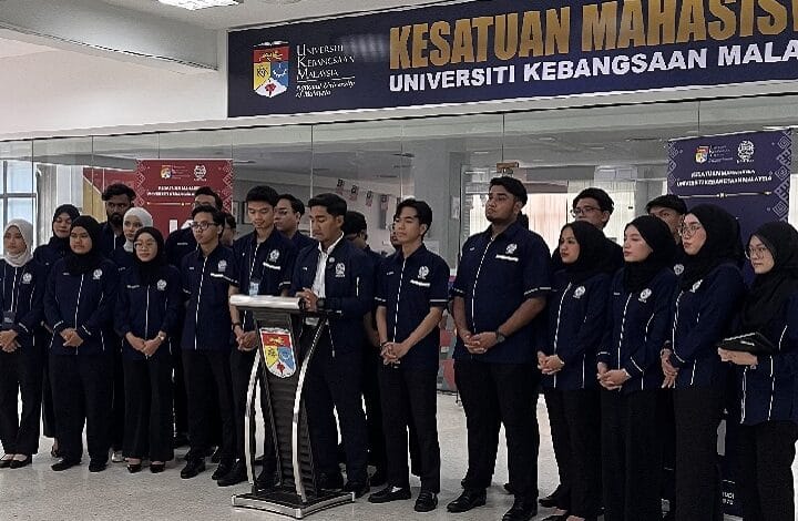 Group of students in navy uniforms stand behind a podium in a university lobby; banner overhead reads 'KESATUAN MAHASISWA' and 'UNIVERSITI KEBANGSAAN MALAYSIA'.