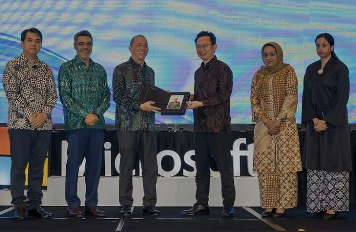 Six people in batik attire stand on a stage as two men exchange a framed award photo; backdrop displays blue-purple light pattern and a banner behind them.