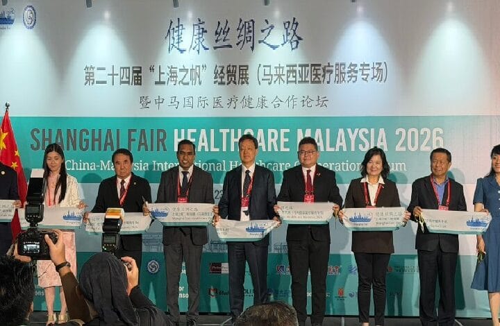 Eight officials in business attire stand on a stage holding large placards during the Shanghai Fair Healthcare Malaysia 2026 ceremony, with a banner in Chinese and English behind them.