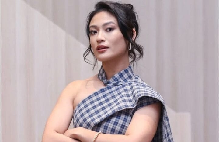 Woman in a blue plaid one-shoulder top posing confidently, arms crossed, neutral backdrop.