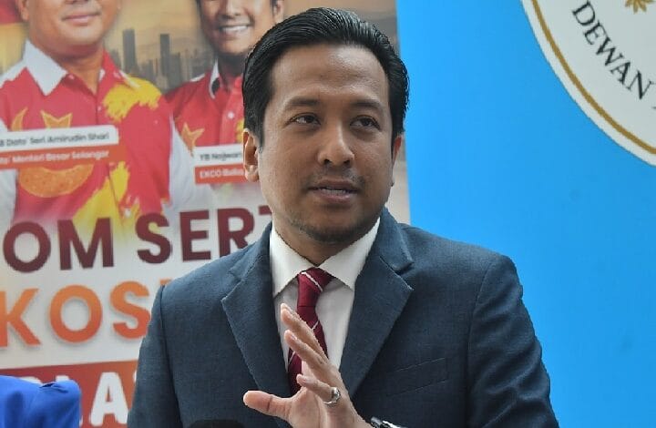 Man in a navy suit and red tie gestures with his right hand while speaking at an event, with a blue wall behind and a poster visible behind him.