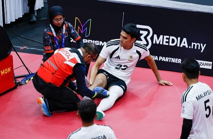 Futsal player #23 in white sits on a red mat as a medic tends to his injured ankle; teammate #5 watches nearby.