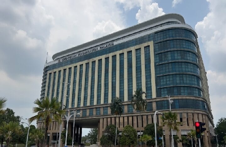 Modern government office building in Malaysia labeled 'Kementerian Pengangkutan Malaysia' with tall glass windows and palm trees in front, daytime setting