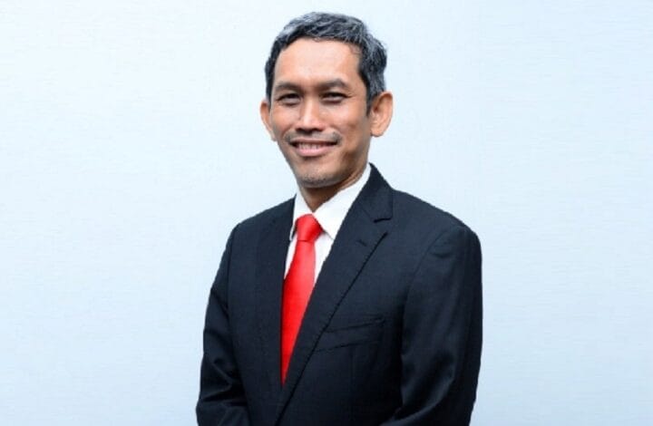 Smiling man in a dark suit with a red tie against a light blue background.