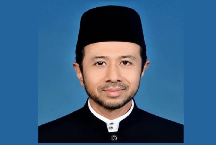 Portrait of a man in a black traditional cap and matching high-collar outfit against a blue backdrop, facing the camera with a subtle smile.