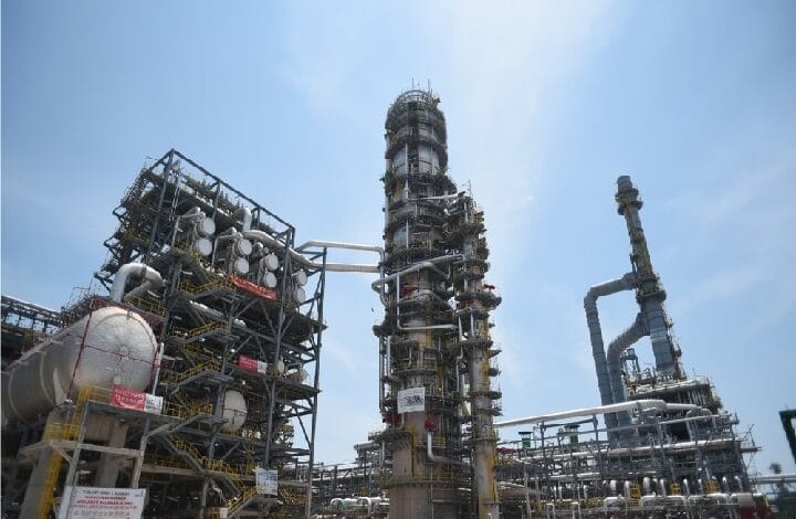 Industrial refinery facility with a tall distillation column, network of pipes, tanks, and catwalks against a clear blue sky.