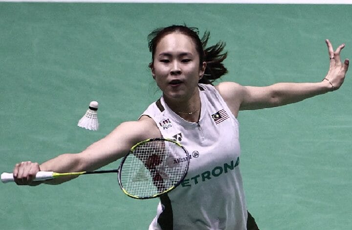 Female badminton player lunges to hit a shuttlecock mid-air on a green court, racket pointed forward and left arm extended for balance.