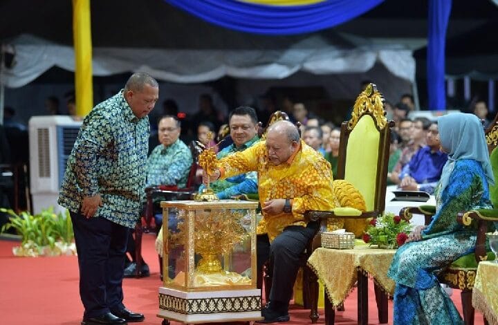 Man in a yellow batik shirt places a ceremonial token into a gilded glass case while suited attendants and a seated official observe a formal ceremony on a red carpet.