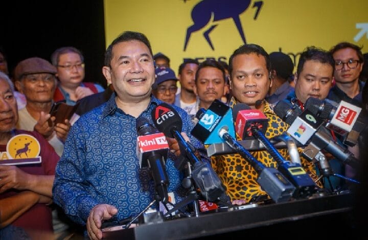 Two men speak at a crowded press conference, standing behind a podium with many microphones while reporters and the audience watch, yellow banner with a logo in the background.