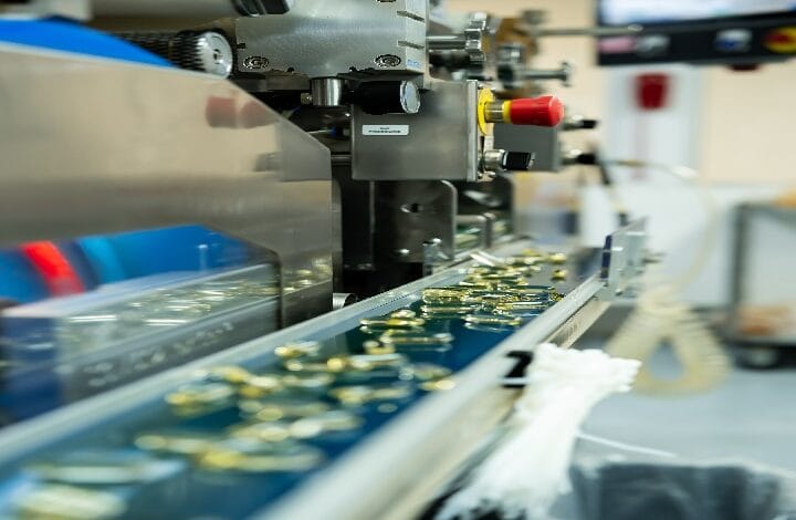 Industrial coin handling machine: coins move along a conveyor belt beneath metal tooling, with a red emergency stop visible in the background.