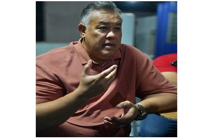 Middle-aged man in a pink polo shirt gesturing with his hands while speaking in a discussion setting.