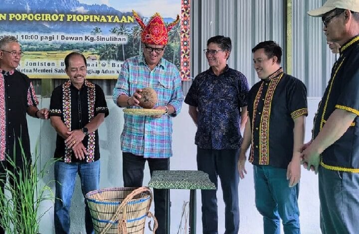 Six men stand at a cultural event; the central man wearing a colorful hat presents a coconut on a tray while others watch and smile.