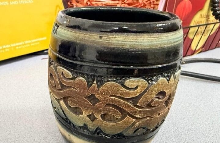 Decorative dark ceramic pot with tan carved scrollwork around the middle, open top, on a kitchen counter with a nearby cord and blurred background objects.