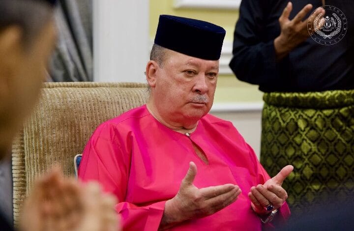 Man in a bright pink traditional robe and black cap sits on a chair, gesturing with hands during a discussion.