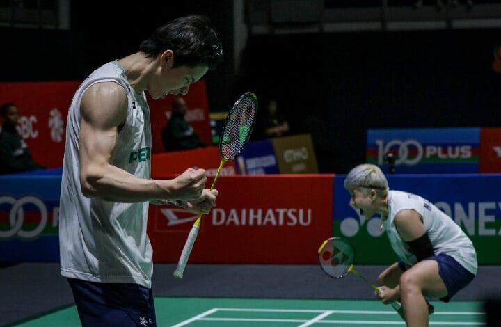 Two badminton players on a court celebrate with rackets in hand; the man in a sleeveless shirt flexes his arm as the woman crouches in the background.