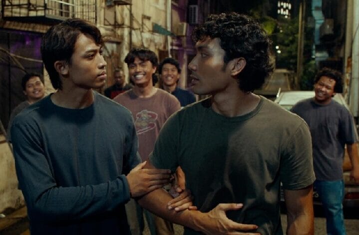 Two men in a nighttime urban alley walk together; one grips the other's arm as they talk, with a group of smiling friends behind them.
