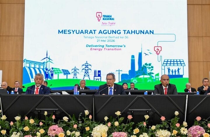 Panel of officials seated at a long table on a stage with a large banner behind reading 'MESYUARAT AGUNG TAHUNAN' and 'Delivering Tomorrow's Energy Transition', featuring blue-green skyline illustrations and the Tenaga Nasional Berhad logo; flowers line the front of the stage.