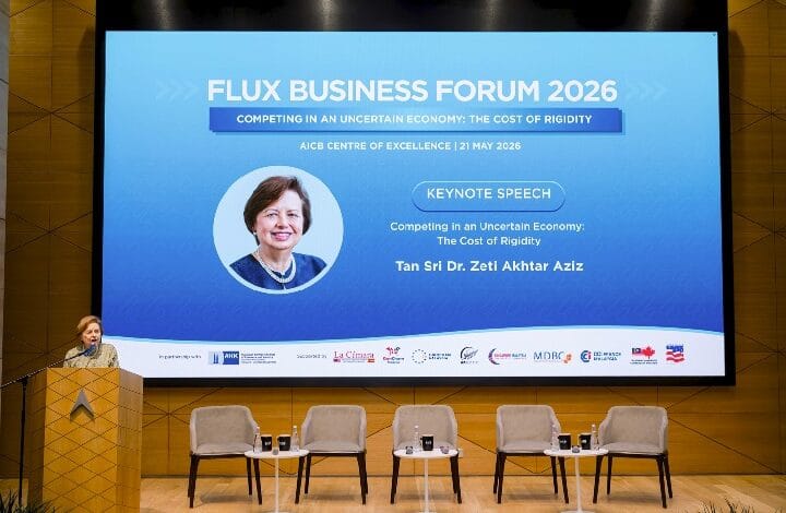 Speaker at podium on a stage with a large blue conference screen reading 'FLUX BUSINESS FORUM 2026' and 'Keynote Speech' (photo of Tan Sri Dr. Zeti Akhtar Aziz shown)