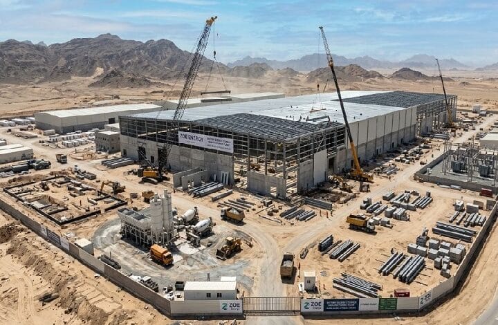 A large industrial warehouse under construction in a desert landscape, with two tall cranes and scattered building materials around the site.